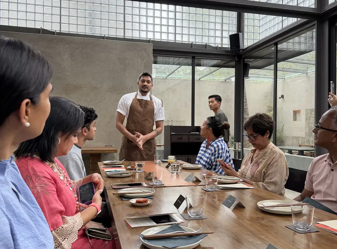A chef in an apron talking to guests seated along a long table