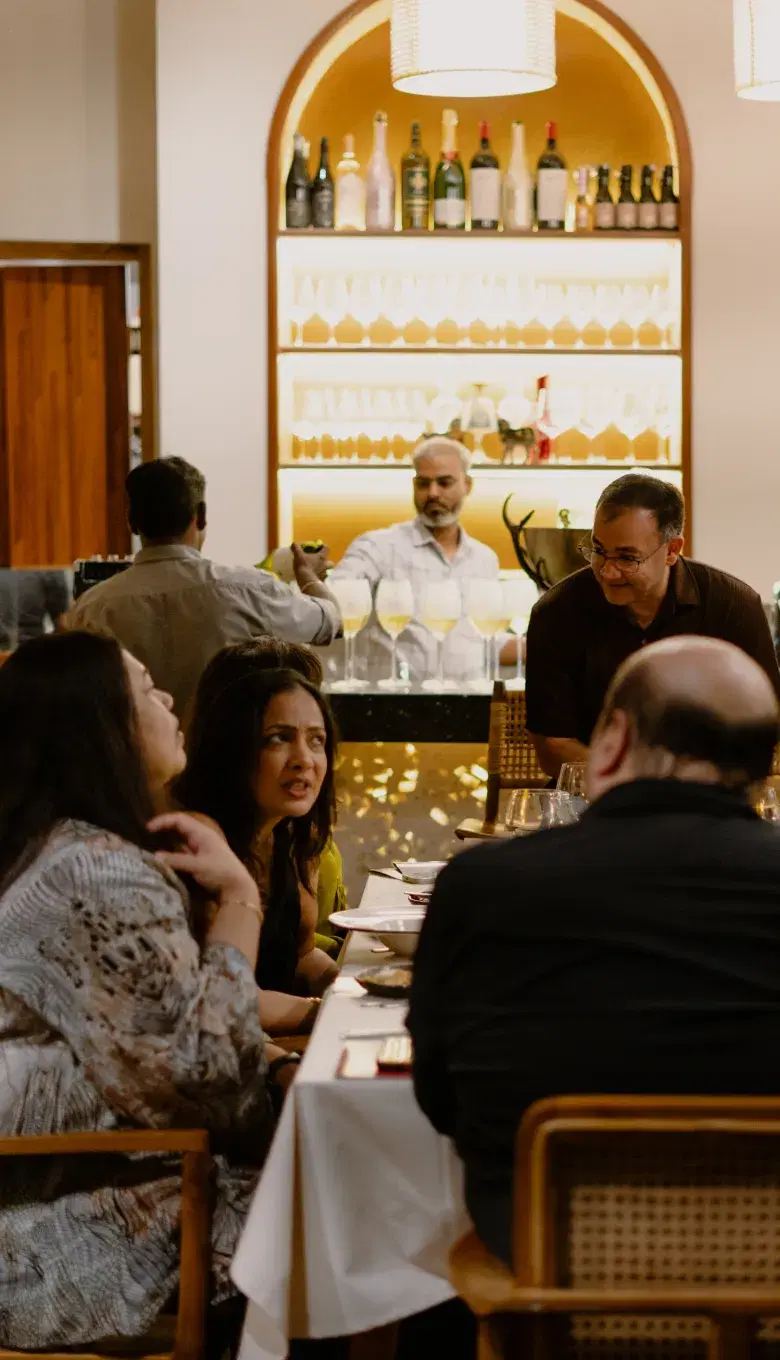 Members at a long dinner table in a restaurant, a lit bar behind them